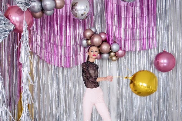 Dancing crazy Fashion girl with beauty makeup, in soft pink pants, mesh bodysuit among confetti, silver shiny foil tinsel. Models having fun with glitter balloons. New Year party. Masquerade party