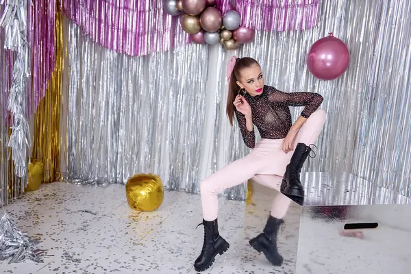 Crazy Fashion girl with beauty makeup, in soft pink pants, mesh bodysuit sits on white cube among confetti, silver shiny foil tinsel. Flexible model fools around with glitter balloons. New Year party