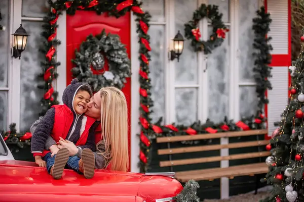 Kafkasyalı annesi olan küçük Afrikalı Amerikalı çocuk Noel arabasının kaputunda oturuyor. Sarışın bir kadının sarıldığı çocuk, evin kırmızı-beyaz kapısının yanında şenlikli bir çelenkle oynaşıyor.