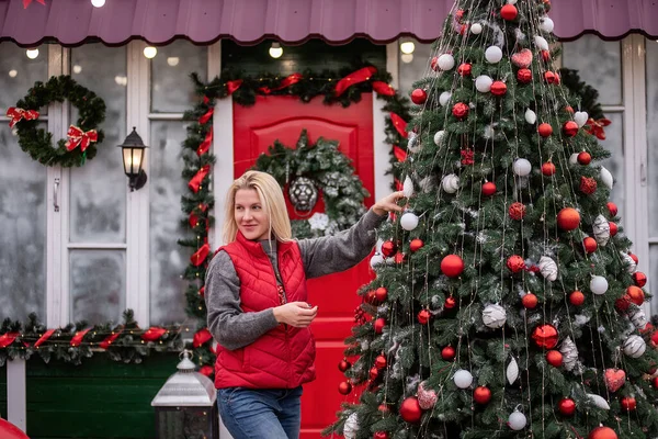 Young blonde woman hangs red balls on Christmas tree on street. Happy girl laughing in puffy vest in backyard decorating exterior of house for New Year. Windows, doors covered with snow, wreath hangs