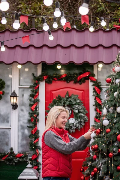 Young blonde woman hangs red balls on Christmas tree on street. Happy girl laughing in puffy vest in backyard decorating exterior of house for New Year. Windows, doors covered with snow, wreath hangs