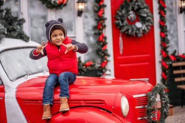 Noel arabasının kaputunda oturan küçük Afrikalı Amerikalı çocuk. Kırmızı beyaz evin kapısında çelenk dallarıyla gezen bir çocuk gülümsüyor. İyi tatiller. Boşluğu kopyala