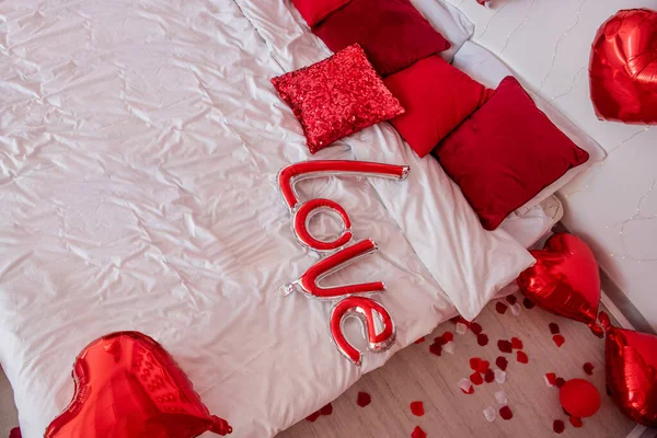 Top view of balloon in form of inscription Love lies on white bed among red pillows. Surprise for Valentines Day. Rose petals on floor. Place for text, copy space, mock up. Design for engagement, date