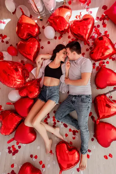 看到一对快乐的年轻夫妇躺在地板上 红白气球心形 玫瑰花瓣四散 情人节浪漫的装饰 男人和女人面带微笑地看着对方 — 图库照片