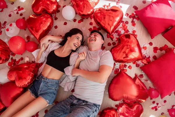 在情人节 这对笑夫妻躺在地板上 玩红箔气球的镜头尽收眼底 年轻的男女拥抱在一起 彼此凝视着被浪漫的装饰包围的玫瑰花瓣 — 图库照片