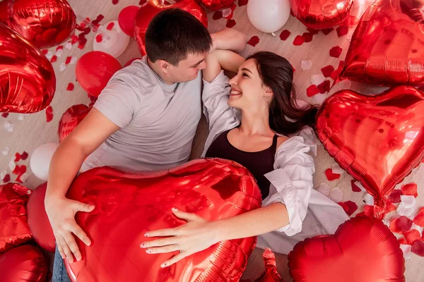 在情人节 这对笑夫妻躺在地板上 玩红箔气球的镜头尽收眼底 年轻的男女拥抱在一起 彼此凝视着被浪漫的装饰包围的玫瑰花瓣 — 图库照片