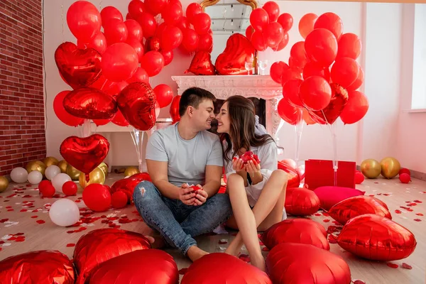 年轻男女坐在靠近白色装饰壁炉的地方 旁边点缀着红球形的心 为庆祝情人节 这对快乐的夫妇捧着一束花瓣欢度浪漫的情人节 — 图库照片