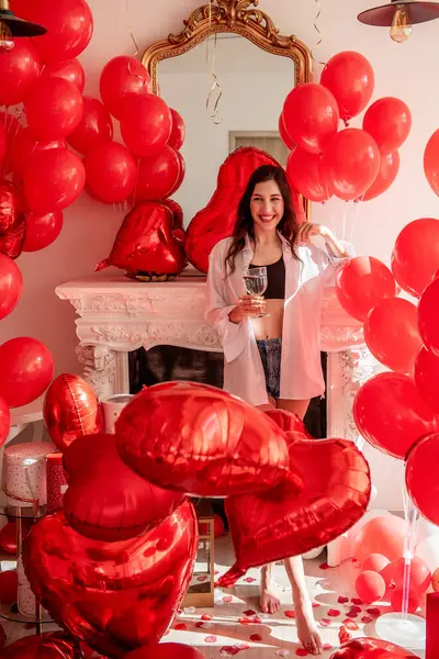 Young woman standing in room filled with mix of red balloons of various shapes, with a romantic white fireplace in the background. Girl holding glass of champagne, joyous smile on face. Valentines day