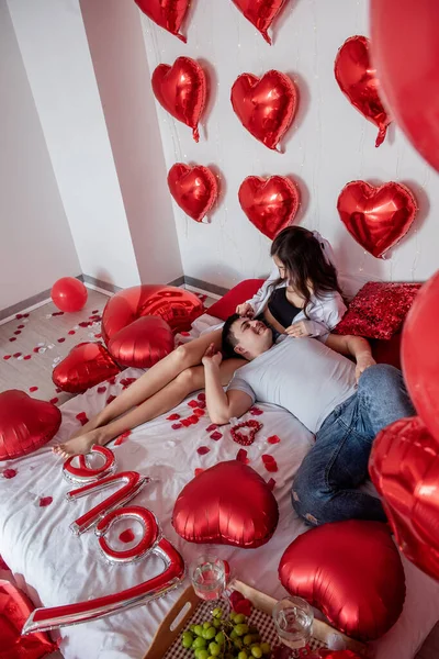 Genç çift rahat ve romantik bir şekilde yatakta dinleniyor. İçi kırmızı kalp balonlarıyla süslenmiş, beyaz duvarda. Kadın oturuyor, sırıtıyor. Sırtüstü uzanmış, başı kucağında.