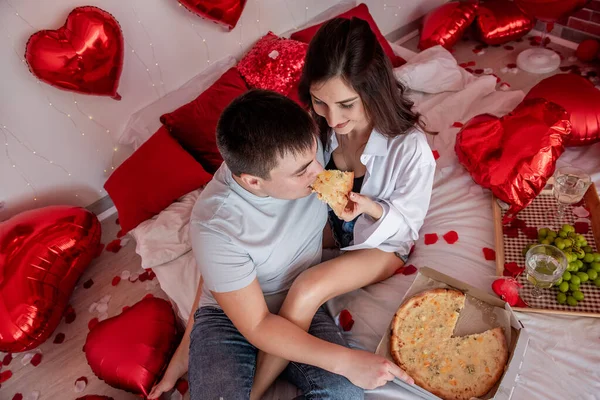 情人节当天 这对快乐的情侣坐在红心气球中的白床上 彼此热恋着 彼此吃披萨 在家里和快餐约会 男人和女人都很开心 — 图库照片