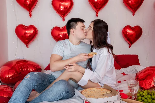 年轻夫妇亲密地在床上亲吻着红心气球 男人和女人都喜欢从外卖盒里拿披萨 喝香槟和葡萄 吃浪漫的饭菜 温馨情人节 — 图库照片