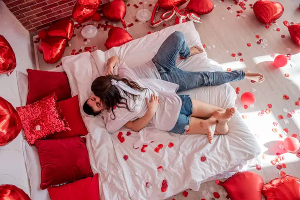 在红球 玫瑰花瓣之间 爱恋的情侣紧紧地拥抱在白床上 男人和女人互相望着对方 在轻松的气氛中度过情人节 惊喜礼物 生活方式 — 图库照片