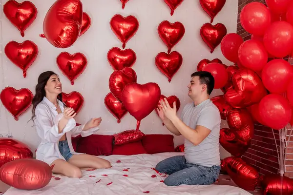 夫妻二人抱着枕头气球在床上打斗 背景是心形红色气球 女人快乐地和男人鬼混 床上撒满了浪漫的玫瑰花瓣 — 图库照片