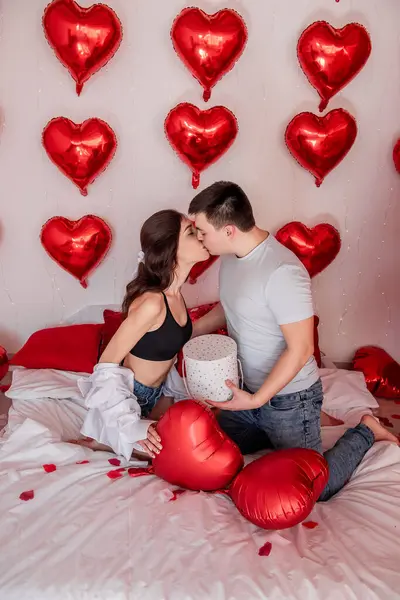情侣们互相亲吻 坐在床上 用心形气球装饰 玫瑰花瓣 庆祝情人节 男人和女人互赠礼物 喜庆的家庭内部 生活方式 — 图库照片