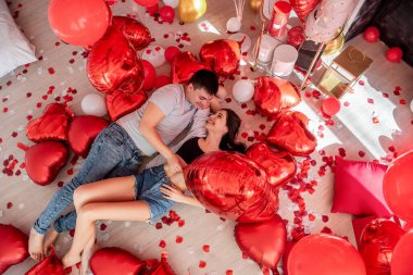 Kırmızı beyaz balon kalp şeklinde yerde yatan, etrafa dağılmış gül yaprakları olan neşeli genç çiftin üst görüntüsü. Sevgililer Günü romantik dekorasyonları. Kadın ve erkek birbirlerine bakıp gülümsüyorlar.