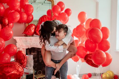 Bir çift oynaşıyor, gülüyor, birbirlerinin gözlerinin içine bakıyor. Erkeklerin sırtında oturan kadın. Hayat tarzı Sevgililer Günü. Beyaz iç mekânlı kırmızı balonların yanında neşeli, romantik bir an.