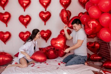 Yatakta kavga eden yastık balonları ve arka planda kalp şeklinde kırmızı balonlar olan çiftler arasında neşeli bir an. Kadınlar neşeyle erkeklerle oynaşıyor. Yatağa romantik gül yaprakları serpilmiş.