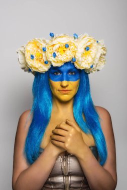 Sarı mavi yüzlü genç bir kadın kafasında şakayık çelengi izole edilmiş gri bir arka planda kurtuluş için dua ediyor. Ukrayna 'da bayrak günü için yaratıcı bir konsept. Uzayı kopyala Kız kameraya bakıyor.