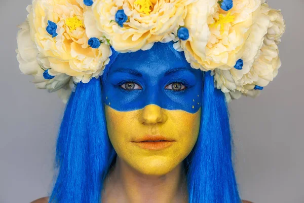 Creative close-up portrait of a young woman with yellow and blue face art, a wreath of peonies on her head on an isolated gray background. Concept for Flag Day in Ukraine. Copy space