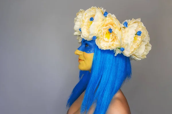 Side view Creative close-up portrait of a young woman with yellow and blue face art, a wreath of peonies on her head on an isolated gray background. Concept for Flag Day in Ukraine. Copy space
