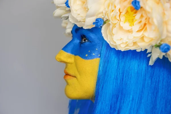 Side view Creative close-up portrait of a young woman with yellow and blue face art, a wreath of peonies on her head on an isolated gray background. Concept for Flag Day in Ukraine. Copy space