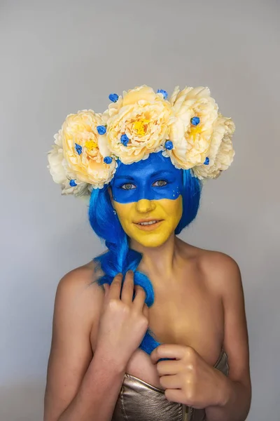 Creative close-up portrait of young happy Ukrainian patriotic woman with yellow blue face art, wreath of flowers on head, holding braid in hands on isolated gray background. Flag Day, Independence Day
