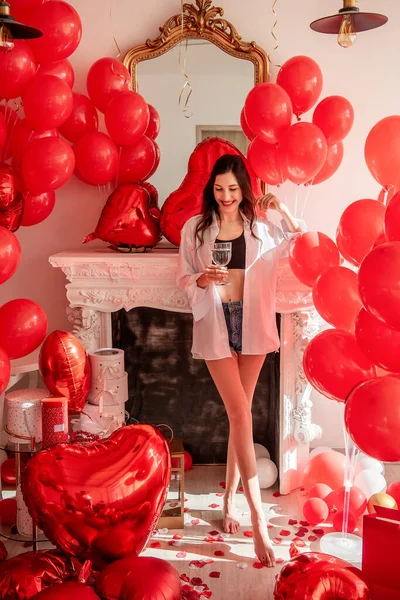 Young woman standing in room filled with mix of red balloons of various shapes, with a romantic white fireplace in the background. Girl holding glass of champagne, joyous smile on face. Valentines day