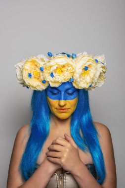 Sarı yüzlü genç bir kadın, kafasında şakayık çelengi, el ele tutuşmuş, gri arka planda kurtuluş için dua ediyor. Ukrayna 'da bayrak günü için yaratıcı bir konsept. Portre yakın plan