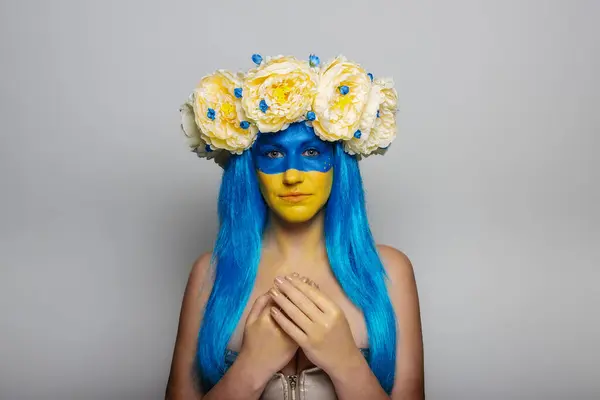 Young woman with yellow blue face art, wreath of peonies on head, praying for salvation on an isolated gray background. Creative concept for Flag Day in Ukraine. Copy space. Girl looking at camera