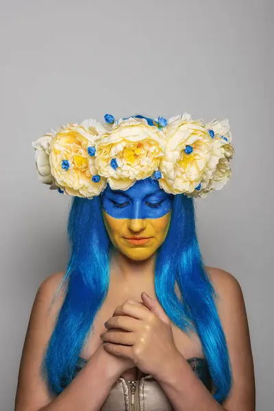 Young woman with yellow blue face art, wreath of peonies on head, holding hands to chest, praying for salvation on isolated gray background. Creative concept for Flag Day in Ukraine. Portrait close-up