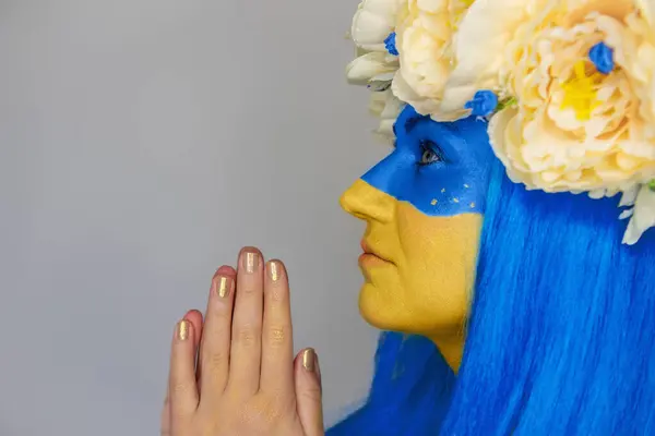 Young woman side view with yellow blue face art, wreath of peonies on head, holding hands to chest, praying for salvation on isolated gray background. Flag Day in Ukraine. Creative portrait close-up