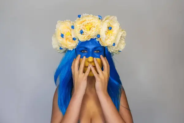 Beautiful young woman covers face with hands in horror. Face of girl in yellow blue color with blue hair, wreath of flowers on head on isolated gray background. Ukrainian Flag Day. Copy space