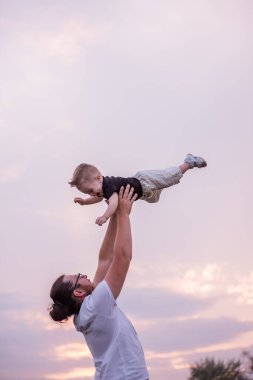 Günbatımında, genç ve çeşitli bir baba, küçük oğlunu yumuşak pastel gökyüzünün zeminine fırlatıyor. Bebekler neşeyle havaya fırlatılır, alacakaranlıkla, neşe ve özgürlüğü simgeleyen.