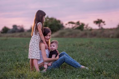 Anne oğlunu ve kızını kucaklıyor, alacakaranlık çökerken çimen tarlasında oturuyor. Huzurlu bir sevgi, bir bağ anı. Dusk 'ta Serene Family Time. Yeşil çayırda çocuklara sarılan bir kadın. Anneler Günü