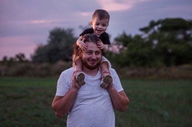 Oğlu omuzlarında taşıyan sakallı bir baba, alacakaranlık aydınlığında, baba sevgisinin özünü somutlaştıran hassas bir anı paylaşıyor. Erkek ve çocuk oynaşıyor, oynuyorlar. Çocuk burnuna dokunur.