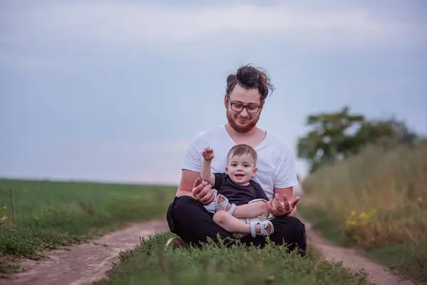 Seated on tranquil path, young, diversity father with happy son on lap, both enjoying peaceful twilight atmosphere in countryside. Man with hair bun, beard playing with funny boy