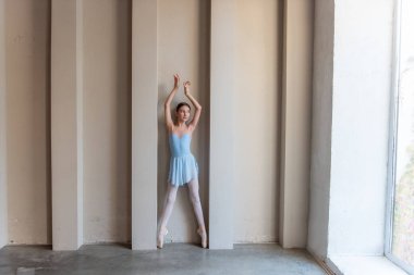 Mavi kostümlü zarif genç balerin, kollarını nazikçe kaldırarak sanatın ve yapının kontrastını yaratan, güneş ışığıyla aydınlatılmış mimari özelliklerin önünde dikiliyor. Zarif Bale Pozisyonu