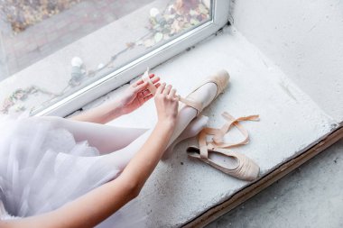 Tepeden bakıldığında, parlak bir pencerenin önünde oturan bir balerin titizlikle dans etmek için sivri uçlu ayakkabılarını bağlıyor, dışarıdaki şehir manzarasıyla çevrili..