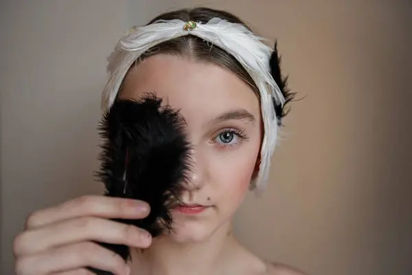 Young ballerina girl with feathered headband tiara holds a black feather over her face, eyes revealing curiosity, hint of playful mischief. Swan Lake concept