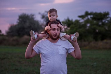 Oğlu omuzlarında taşıyan sakallı bir baba, alacakaranlık aydınlığında, baba sevgisinin özünü somutlaştıran hassas bir anı paylaşıyor. Erkek ve çocuk oynaşıyor, oynuyorlar. Çocuk burnuna dokunur.