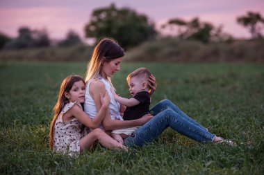 Anne oğlunu ve kızını kucaklıyor, alacakaranlık çökerken çimen tarlasında oturuyor. Huzurlu bir sevgi, bir bağ anı. Dusk 'ta Serene Family Time. Yeşil çayırda çocuklara sarılan bir kadın. Anneler Günü