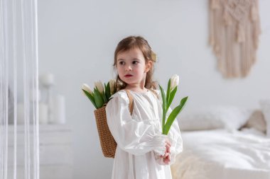 Close-up portrait of a little girl in a white dress, nightgown. Toddler embraces a bouquet of fresh, delicate white tulips. Gift for the holiday, the concept of purity, spring time. Copy space
