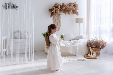 Beyaz elbiseli küçük bir kız elinde lalelerle bohem bir yatak odasında duruyor. Minimalist odada doğal dekor, yumuşak ışıklandırma, dingin ve rahat bir atmosfer yaratıyor. Pampas çimen Boho iç mimarisi