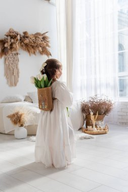 Little girl in white dress, nightgown stands with back, faceless, on shoulders is hand-made backpack made of raffia with tulips. Light airy, spring interior in rustic style. Pampas grass on the wall