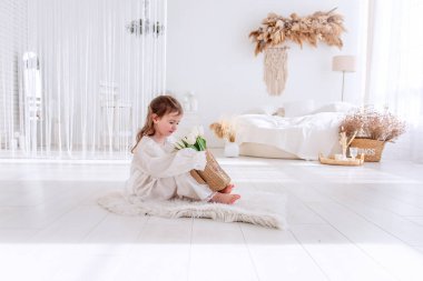Beyaz Boho minimalist iç mekanında küçük bir kız yerde oturuyor. Bebek elbiseli, gecelikli, elinde bir buket bahar lalesi tutan, sarılan, çiçek kokan. Airy, ışık dolu bir yer. Pampas çimen dekoru
