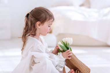 Close-up portrait of a little girl in a white dress, nightgown. Toddler embraces a bouquet of fresh, delicate white tulips. Gift for the holiday, the concept of purity, spring time. Copy space