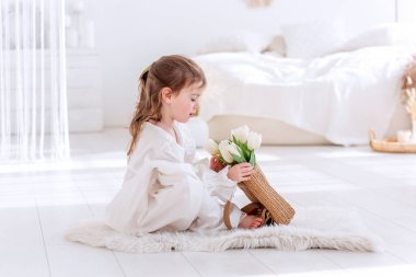 Beyaz Boho minimalist iç mekanında küçük bir kız yerde oturuyor. Bebek elbiseli, gecelikli, elinde bir buket bahar lalesi tutan, sarılan, çiçek kokan. Airy, ışık dolu bir yer. Pampas çimen dekoru