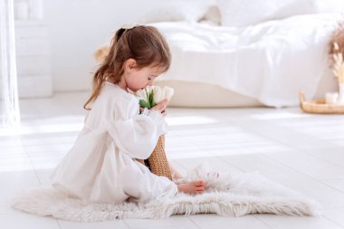 Beyaz Boho minimalist iç mekanında küçük bir kız yerde oturuyor. Bebek elbiseli, gecelikli, elinde bir buket bahar lalesi tutan, sarılan, çiçek kokan. Airy, ışık dolu bir yer. Pampas çimen dekoru