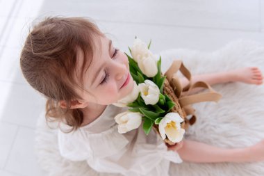 Top view close-up portrait of little girl in a white dress, nightgown. Toddler embraces a bouquet of fresh, delicate white tulips. Gift for the holiday, the concept of purity, spring time. Copy space