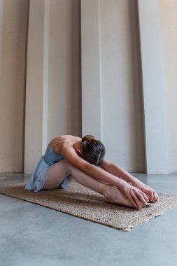 Genç balerin, sakin bir esneme noktasında, bir minderin üzerinde otururken öne doğru eğiliyor, odaklanmayı ve disiplini vurgulayan mimari sütunlarla çevrili..